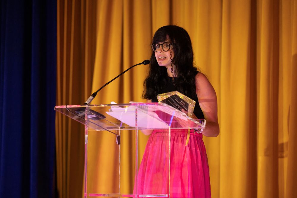 Jess Wade, recipient of the 2024 SPIE Directors' Award, speaks in front of a gold curtain after accepting her award at the 2025 SPIE Society Awards banquet