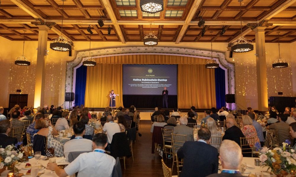 Halina Rubinsztein-Dunlop accepting the Gold Medal on stage at the 2025 SPIE Society Awards banquet, with a large audience seated at round tables in a grand, chandelier-lit room.