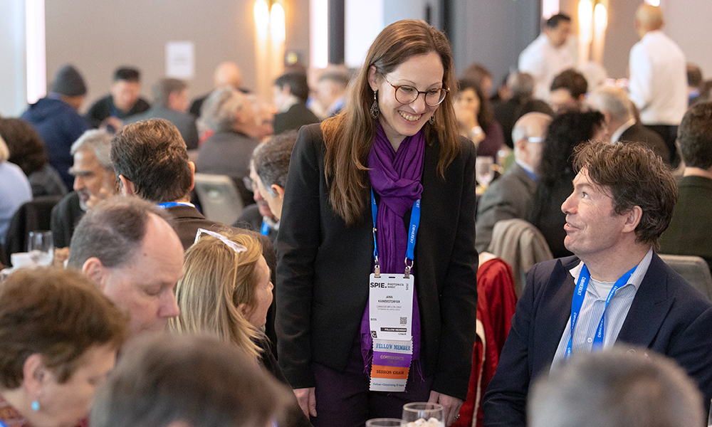 An SPIE Fellow stands and converses with attendees at an SPIE event. The Fellow is wearing a purple scarf and a name badge indicating their status as a Fellow Member. The event appears to be well-attended, with many people seated at tables in the background.