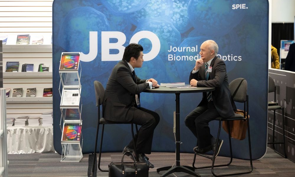 Two individuals in business attire are seated at a high table, conversing at an SPIE event in front of a 'JBO Journal of Biomedical Optics' backdrop, with a brochure stand filled with colorful SPIE Publications and Journals pamphlets nearby.