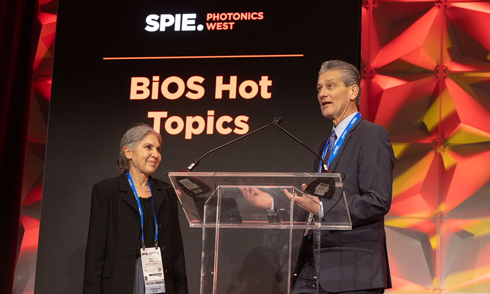 Two individuals stand on stage at BiOS Hot Topics during SPIE Photonics West. They are positioned behind a clear podium with microphones, and the backdrop features the event's name in bold letters along with a geometric design in red and yellow hues.