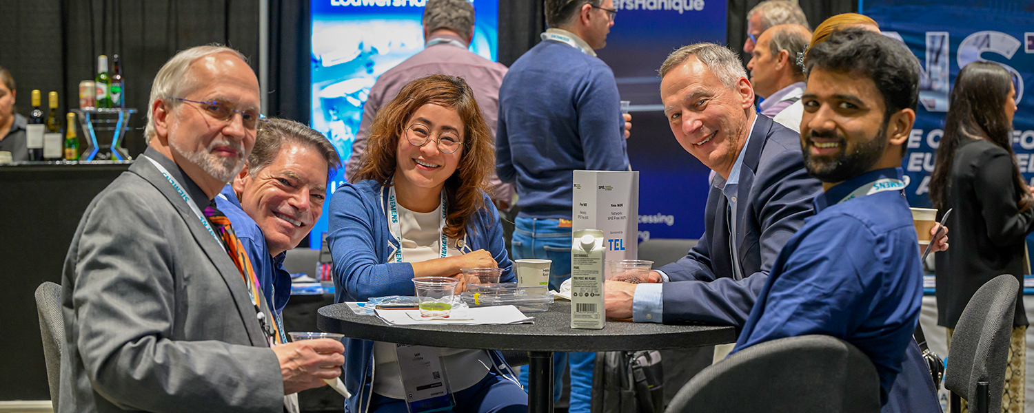 Several attendees at an SPIE meeting sit around a round table, engaged in conversation. The background includes other conference attendees and exhibition booths with various displays and banners.