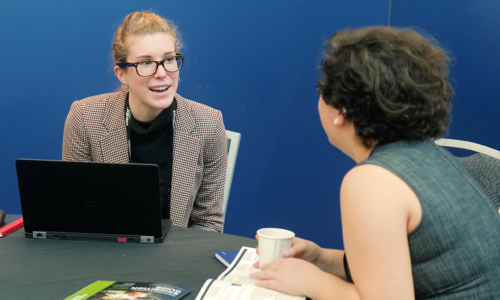 Two people talking at a table, one with a laptop, the other holding a cup and papers, against a blue background