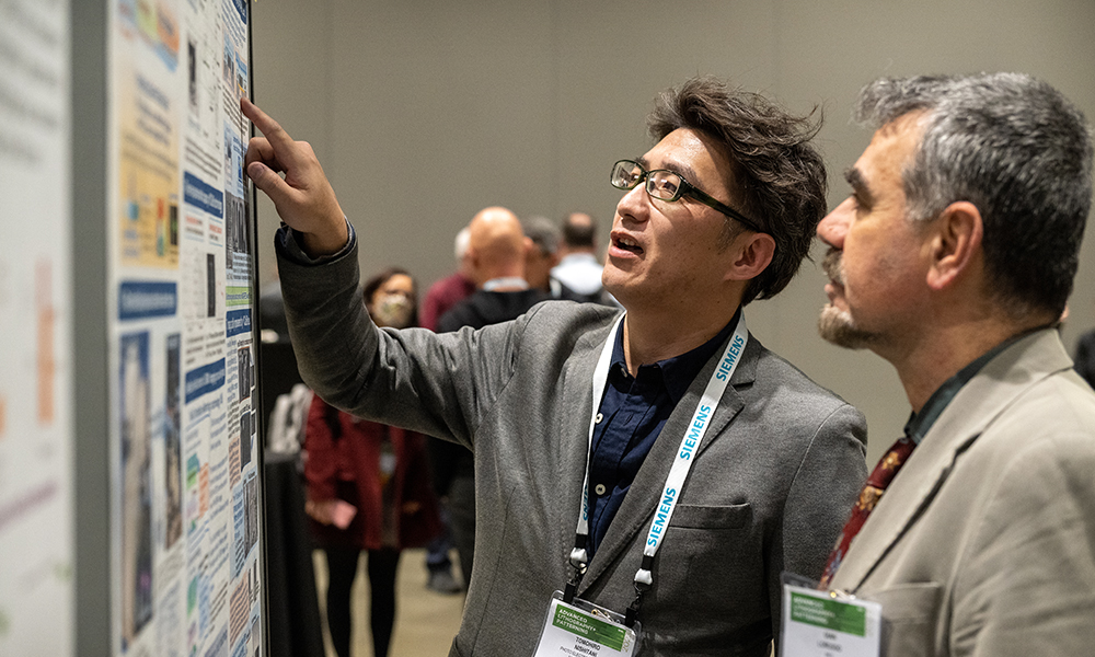 Two people stand closely together at a conference poster session, with one of them pointing to information on a large research poster while discussing its content. Other attendees and posters are visible in the background.