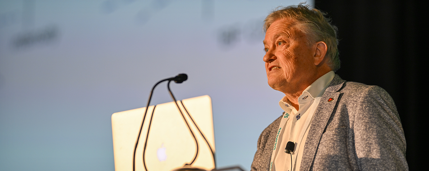Person in a light shirt and gray blazer presenting at a podium with a laptop, in front of a blurred projection screen.
