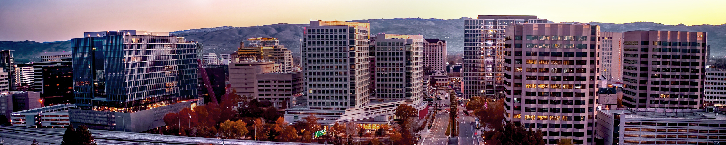Wide panoramic view of downtown San Jose at sunset, featuring glass and concrete office towers, quiet city streets lined with fall foliage, and the Santa Clara Valley hills in the distance.