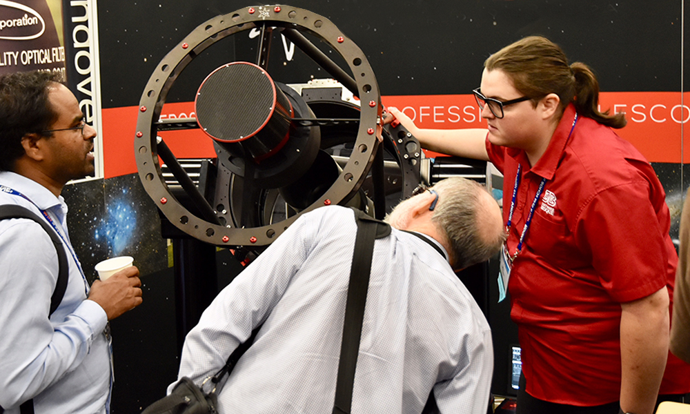 Attendees at an exhibition booth examining a large black telescope mount with a circular frame and central optical component; one person gestures toward the equipment while others observe closely, with astronomy-themed signage in the background.