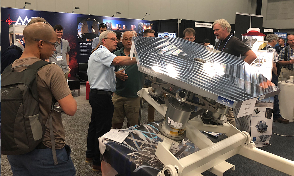 Attendees gathered around a large metallic optical instrument with a reflective, ridged surface mounted on a sturdy base at an exhibition booth; surrounding displays feature technical posters and equipment related to astronomy and optics.