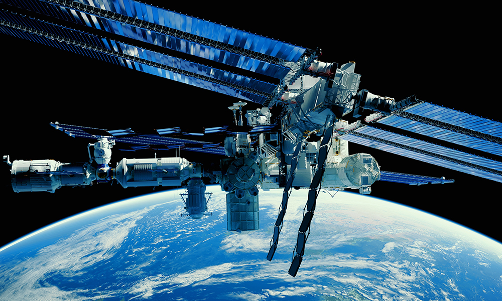 The International Space Station (ISS) orbiting above Earth, with its solar panels fully extended. The station is illuminated by sunlight, set against the dark backdrop of space. Below, Earth’s blue oceans and swirling white clouds are clearly visible.