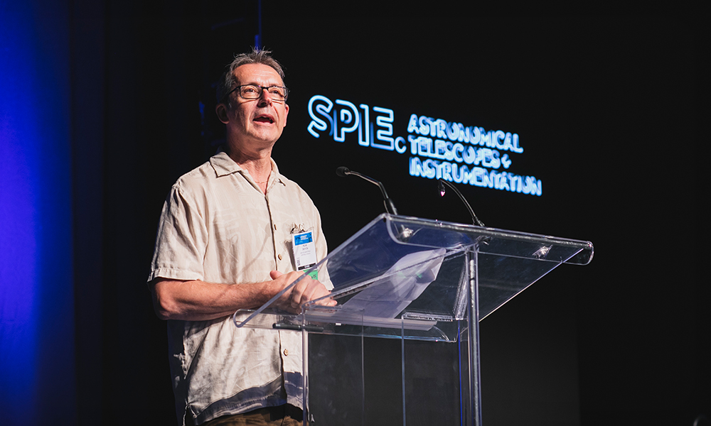 A speaker stands at a clear podium, addressing an audience at the SPIE Astronomical Telescopes + Instrumentation conference. The individual wears glasses, a light-colored shirt, and a name badge, with the event name illuminated in the background.