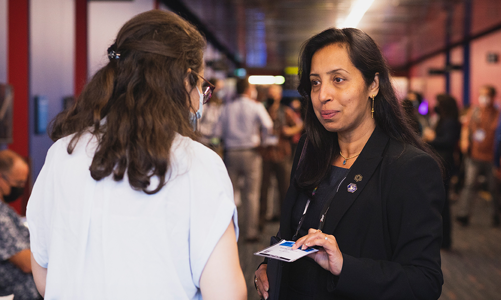 Two individuals engaged in conversation at a professional event. One person, with long brown hair tied back and wearing a white shirt, faces away from the camera. The other, wearing a black blazer and holding an ID badge, faces the camera.