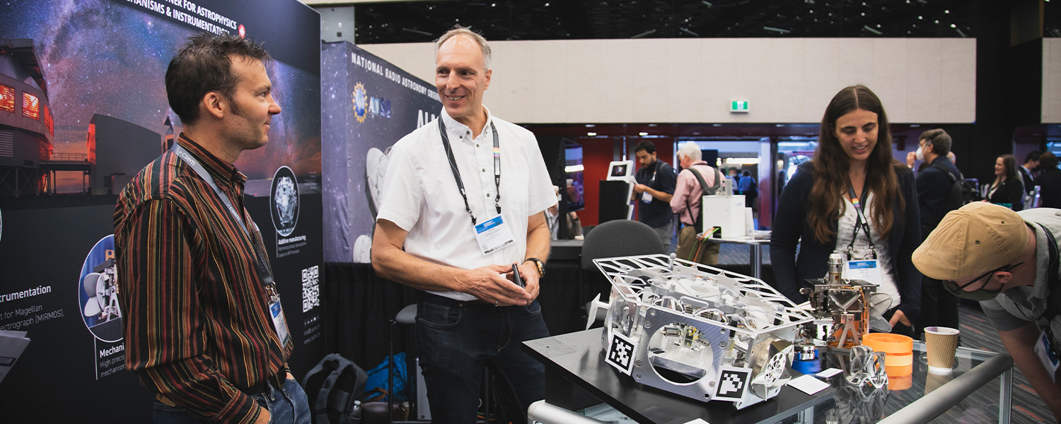 A group of people gather around a table at an telescope exhibition, observing and discussing a detailed mechanical model. The display includes labeled components and is set against a backdrop of posters related to astrophysics and instrumentation. Logos for the National Radio Astronomy Observatory (NRAO) and other scientific organizations are visible.