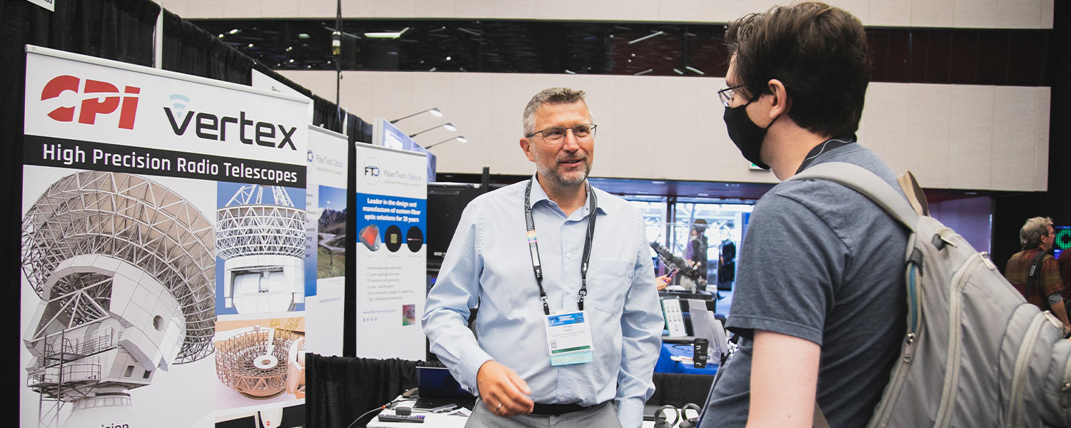 Two attendees at an exhibition booth discussing high precision radio telescopes; a large banner displays the CPI Vertex logo and images of radio telescope structures, with additional informational panels and equipment visible in the background.