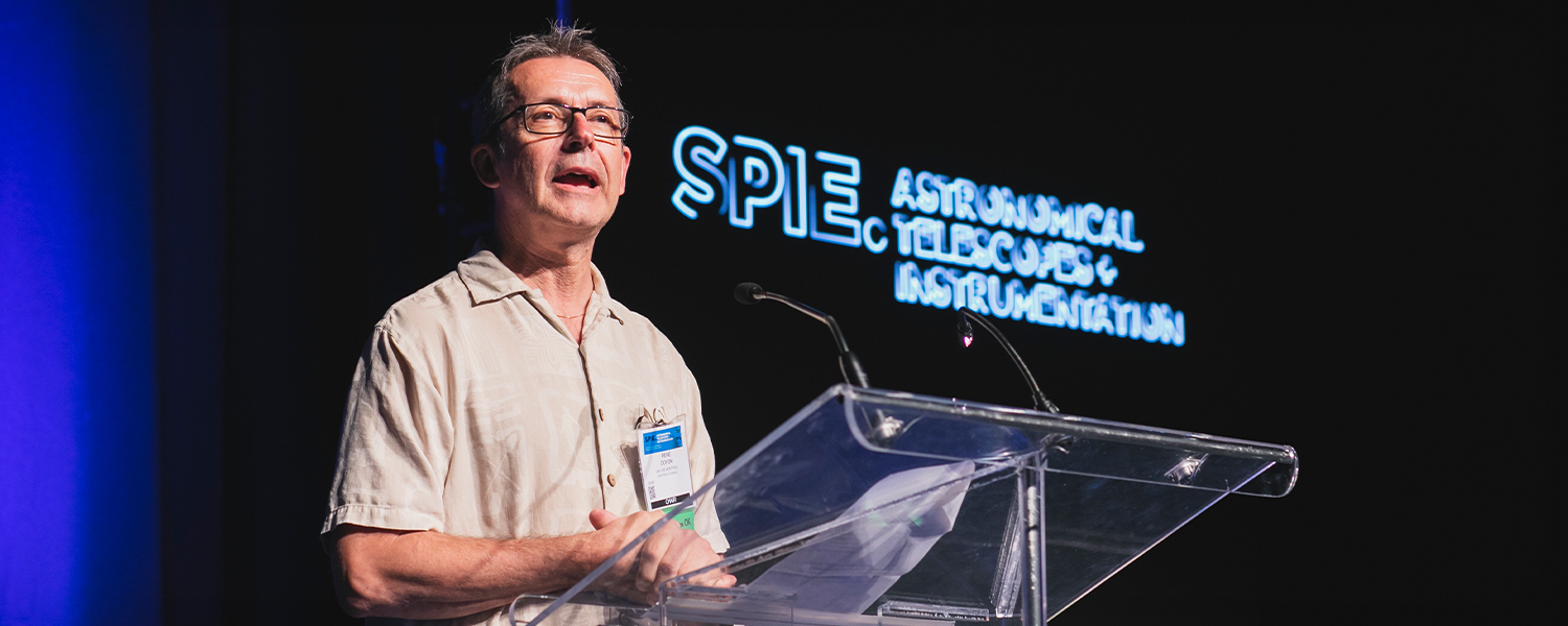 Conference speaker standing at a clear lectern under stage lighting, with SPIE Astronomical Telescopes + Instrumentation signage in the background.