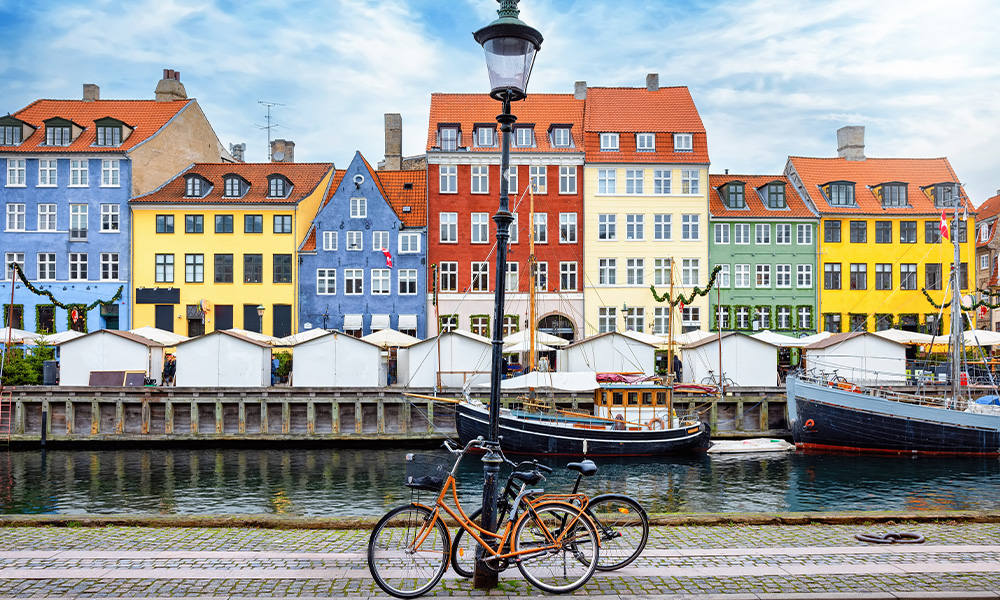 Row of colorful buildings with red, yellow, green, and blue facades along a waterfront canal; boats are docked in the water, and two bicycles are parked near a lamppost in the foreground.