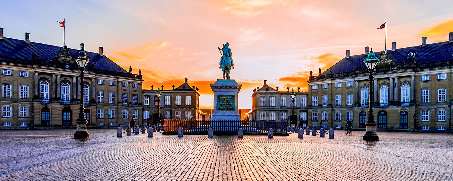 Amalienborg Palace in Copenhagen, Denmark, captured at sunset. Four identical rococo-style palace façades form an octagonal courtyard, with a bronze equestrian statue of King Frederick V at the center. The sky glows in warm shades of orange and pink, casting a golden light over the historic architecture.