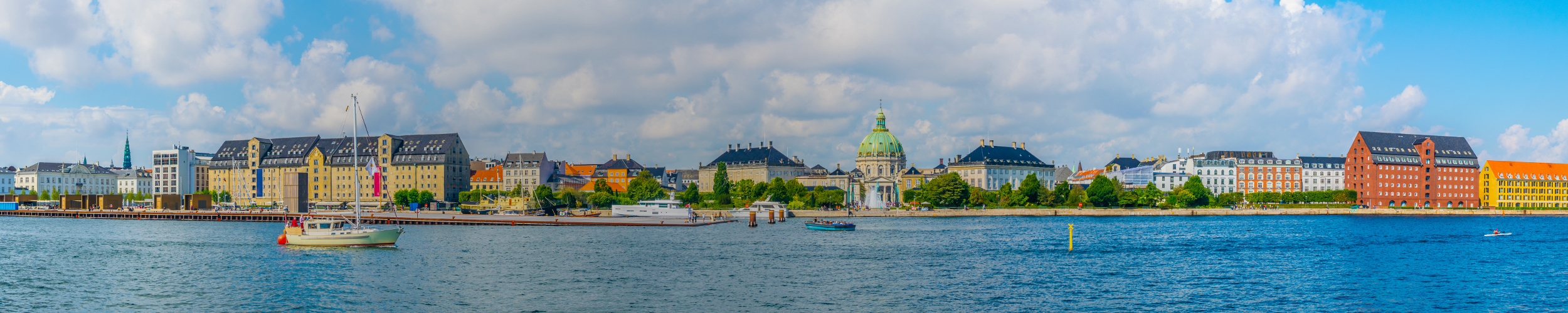 Panoramic view of Copenhagen waterfront with historic buildings, including the dome of Frederik's Church, and boats on the water under a partly cloudy sky. SPIE Astronomical Telescopes + Instrumentation will be hosted in Copenhagen.