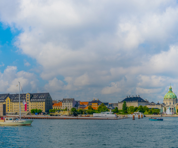 Panoramic view of Copenhagen waterfront with historic buildings, including the dome of Frederik's Church, and boats on the water under a partly cloudy sky. SPIE Astronomical Telescopes + Instrumentation will be hosted in Copenhagen.