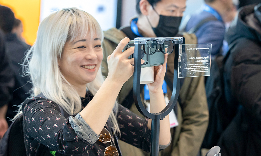 Person with long, light-colored hair holding a camera up to a display of smart glasses mounted on a stand on the show floor of the AR VR MR Expo, with other people visible in the background.