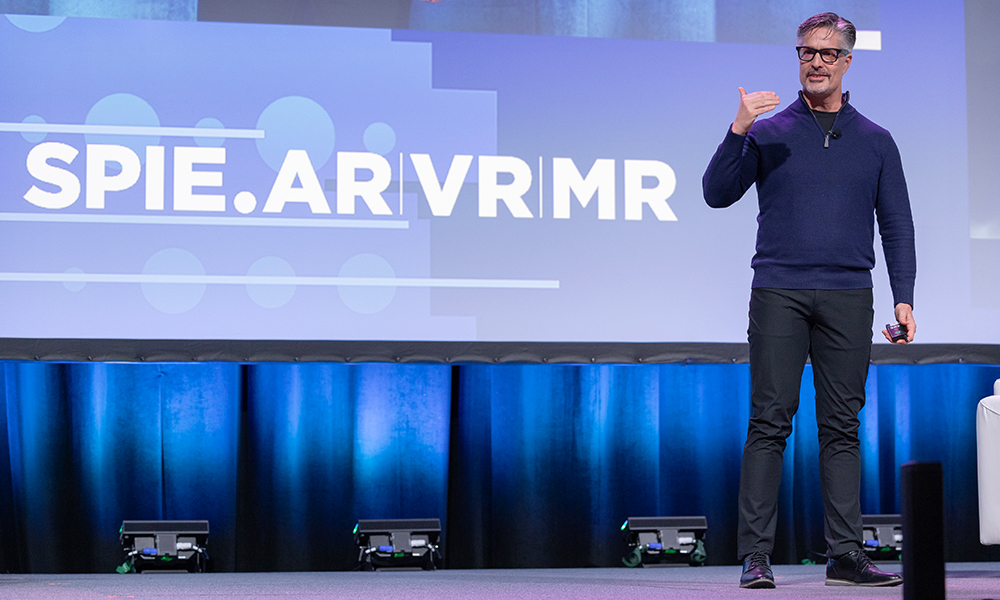 Person standing on stage giving a presentation at SPIE.AR | VR | MR conference, holding a laser pointer in one hand and gesturing with the other hand, with the conference logo and name in the background.