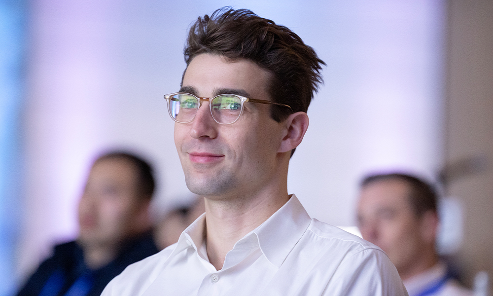 A person with short, dark hair and glasses is wearing a white collared shirt. In the background, there are other people whose faces are blurred, and the setting is indoors with soft lighting and a neutral-colored wall.