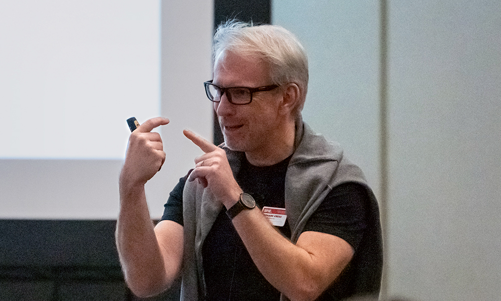 Bernard Kress has short gray hair and glasses. He is a frequent instructor of augmented, virtual, and mix reality topics. He wears a black shirt with a gray sweater draped over his shoulders. He is holding a laser in one hand while gesturing with the other. The background shows part of a projection screen.