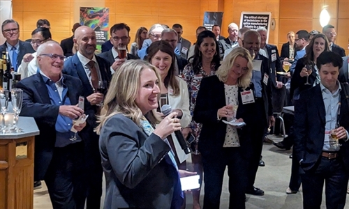 A speaker addresses an audience at a professional indoor event, with attendees in business attire holding drinks and wearing name tags, set in a decorated venue with artwork on the walls.