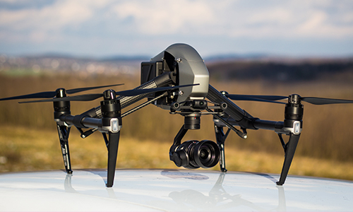 A black and gray quadcopter drone with a mounted camera is resting on a white surface outdoors. The background shows a blurred landscape with fields and hills under a partly cloudy sky.
