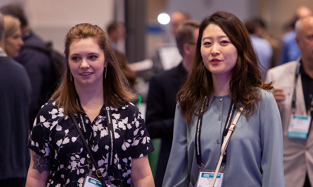 Two individuals wearing conference badges and lanyards walking through the Defense + Commercial Sensing conference with other attendees in the background.