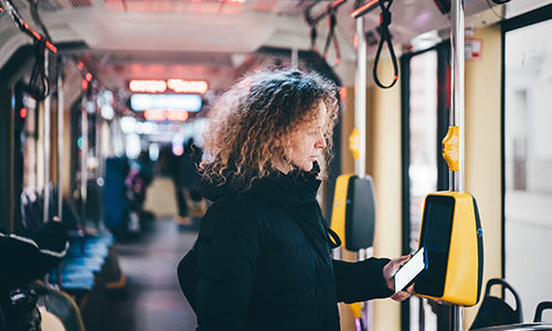 Person standing inside a public transit vehicle, holding a smartphone near a yellow ticket validation machine mounted on a pole. The interior shows rows of empty blue seats, overhead hand straps, and illuminated signage in the background under bright lighting.