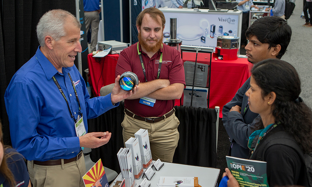 Two people at a SPIE Defense booth demonstrating a large optical lens to a small group, with brochures and event materials on the table.