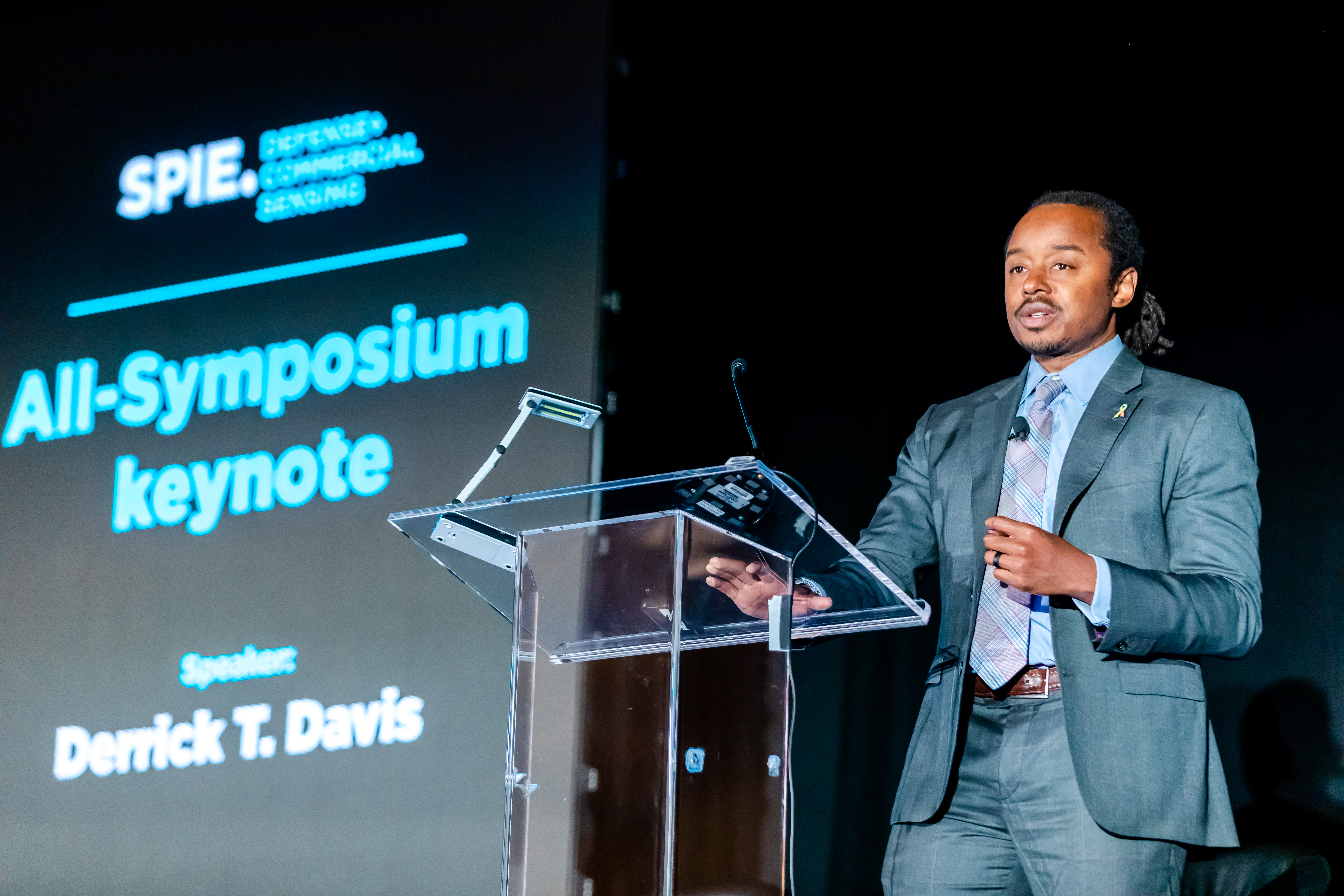 Person in a suit speaking at a podium during an SPIE event, with a screen displaying All-Symposium keynote and the name Derrick T. Davis in the background.