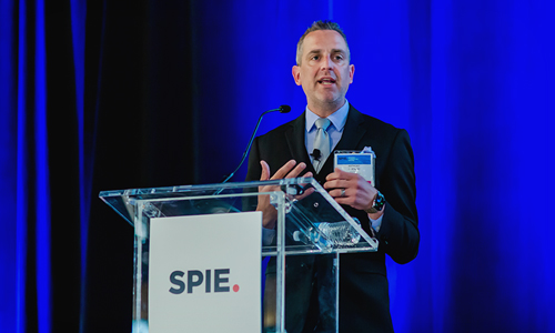 Here’s the ADA-compliant alt text for this image: Alt text: Person standing behind a clear podium with a microphone, speaking at an event with a blue backdrop and a sign displaying the SPIE logo.