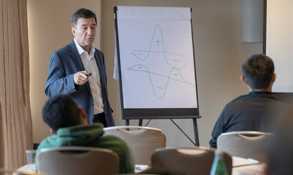 Person in a suit presenting a graph with plus and minus signs on a flip chart to two seated attendees in a conference room.