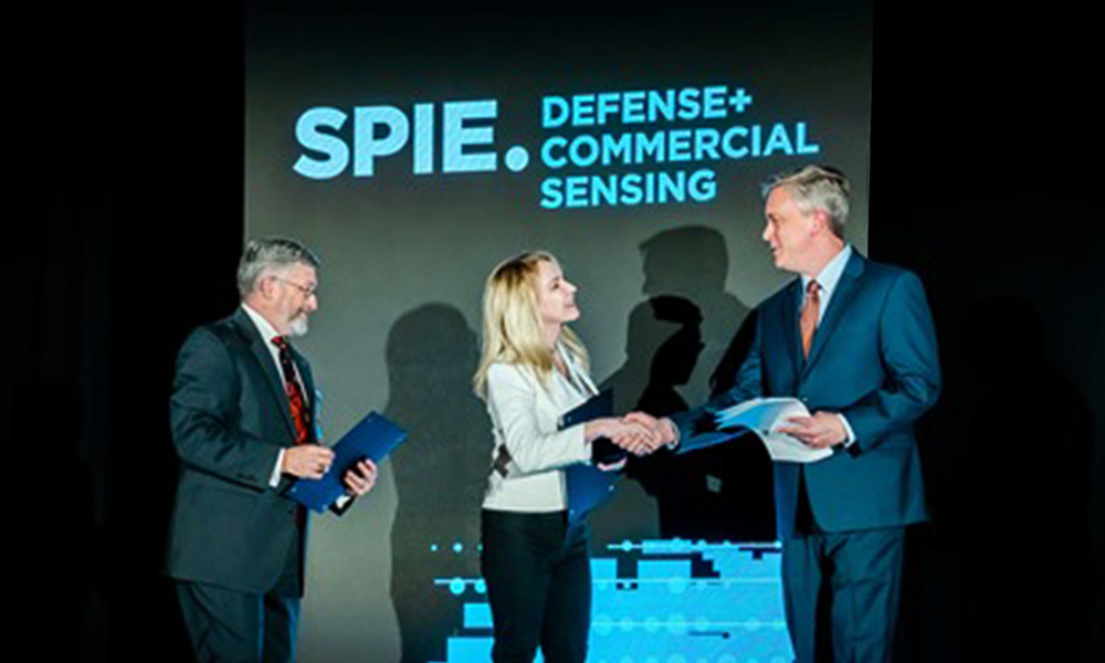 Three individuals on stage at an SPIE Defense and Commercial Sensing event, with two exchanging a handshake and holding documents in front of a large screen displaying the event name.