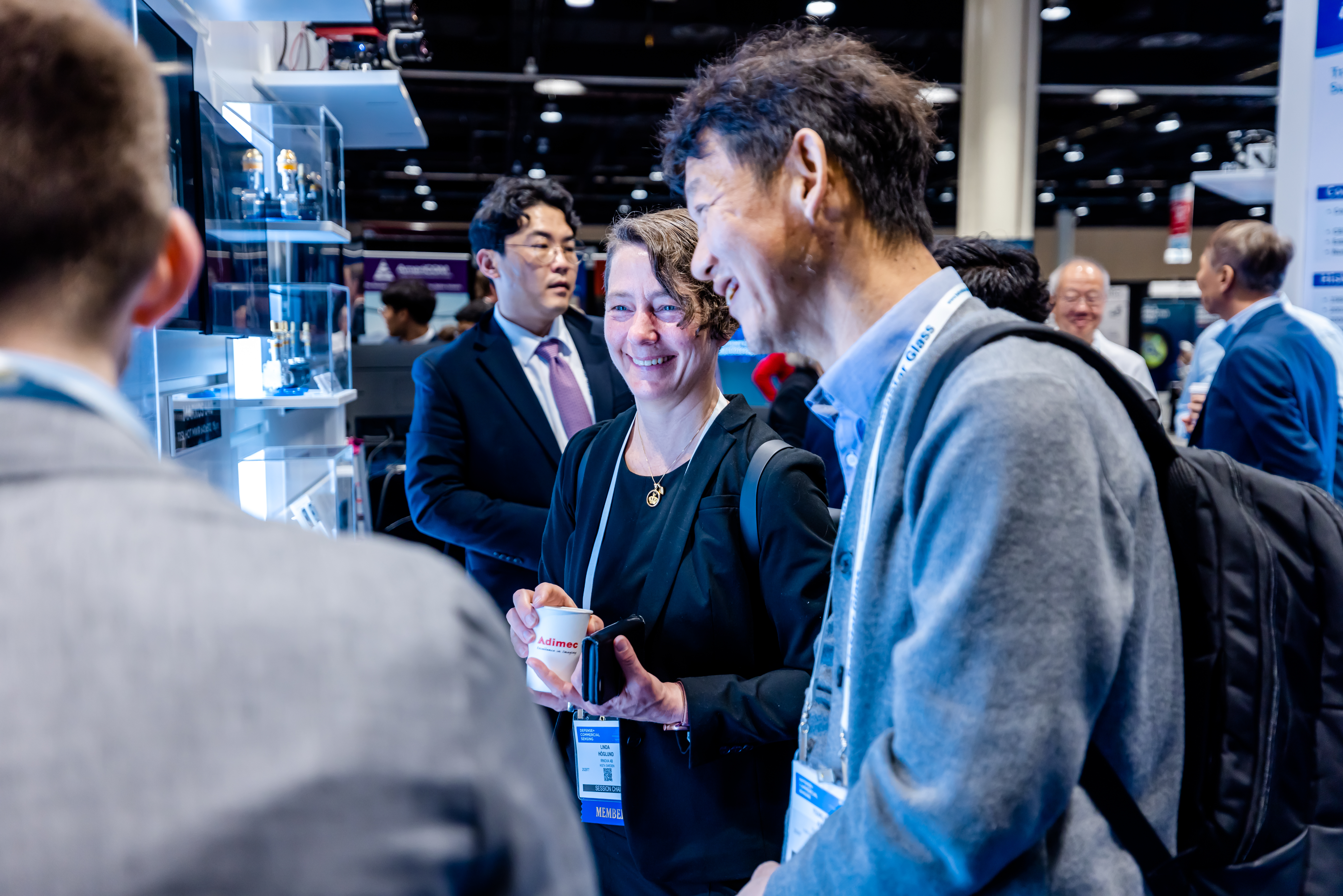 Group of people standing and conversing near a display booth at an indoor event or exhibition.