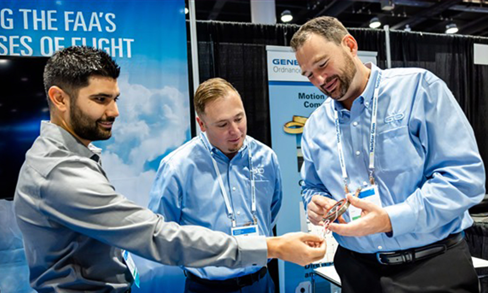 Three individuals are standing at a trade show booth, engaged in a discussion while examining a small object being held by one person. Two of them are wearing matching light blue button-down shirts with lanyards and name badges, while the third person is in a gray shirt. The booth backdrop includes text referencing the FAA and features an image of clouds, with additional signage and equipment visible in the background.