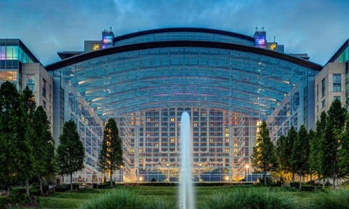 Large glass atrium with a curved roof dominates the center of a modern building, flanked by symmetrical stone and glass wings. A tall water fountain rises in the foreground, surrounded by manicured greenery and trees. The building is illuminated with soft interior lights under a cloudy blue sky.