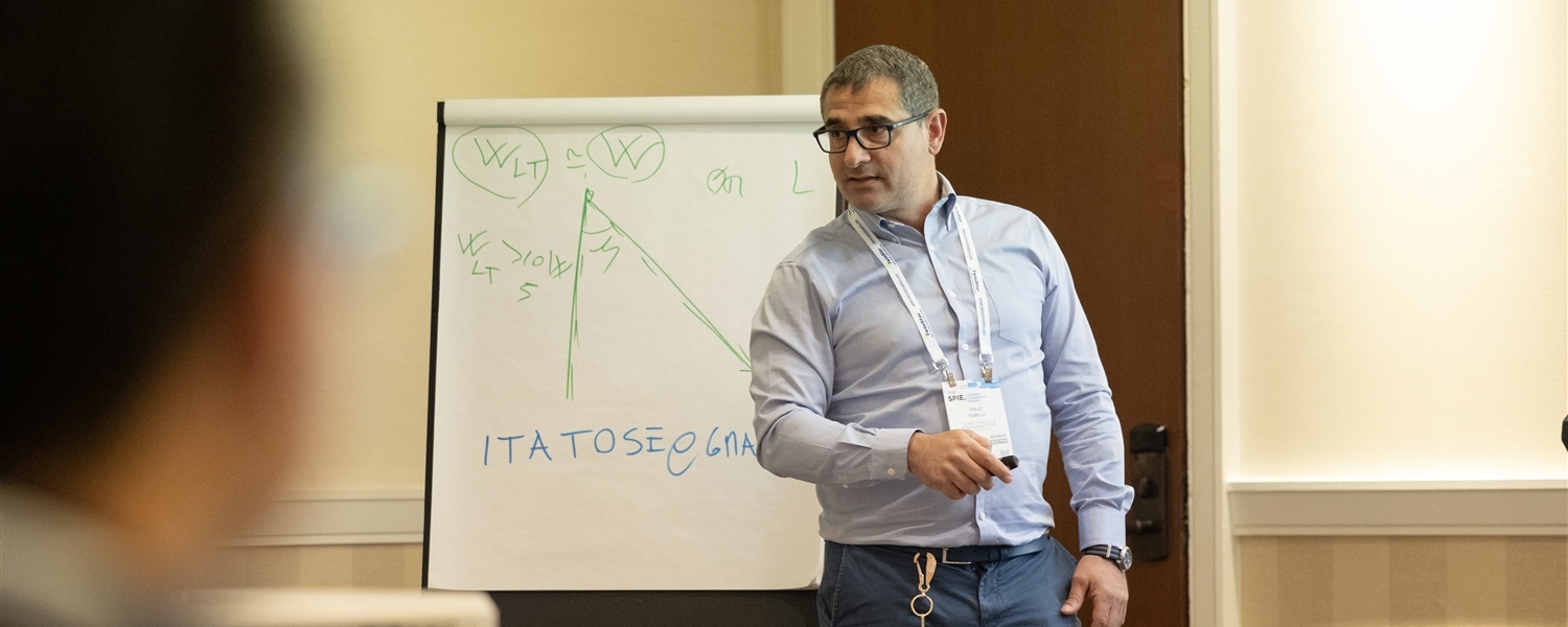 Presenter gestures toward a flip chart showing hand-drawn diagrams and equations during an in-person technical course.