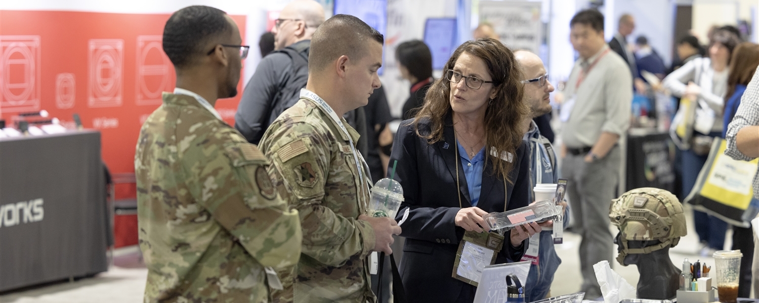 A vendor at a security expo showcases defense technologies to two attendees, who are wearing military outfits.