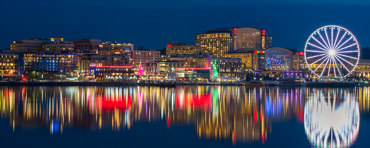 Nighttime view of National Harbor’s waterfront on the Potomac River, with illuminated hotels and shops, festively lit trees along the promenade, and the rainbow-lit Ferris wheel reflecting in the calm water.