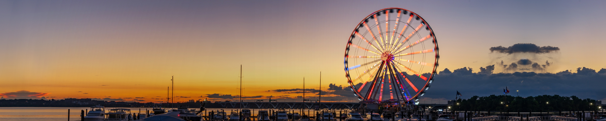 Ferris wheel illuminated with colorful lights at sunset near a waterfront with boats and docks.