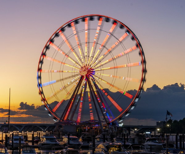 Close-up of a brightly lit Ferris wheel spinning at sunset with boats docked in the foreground.