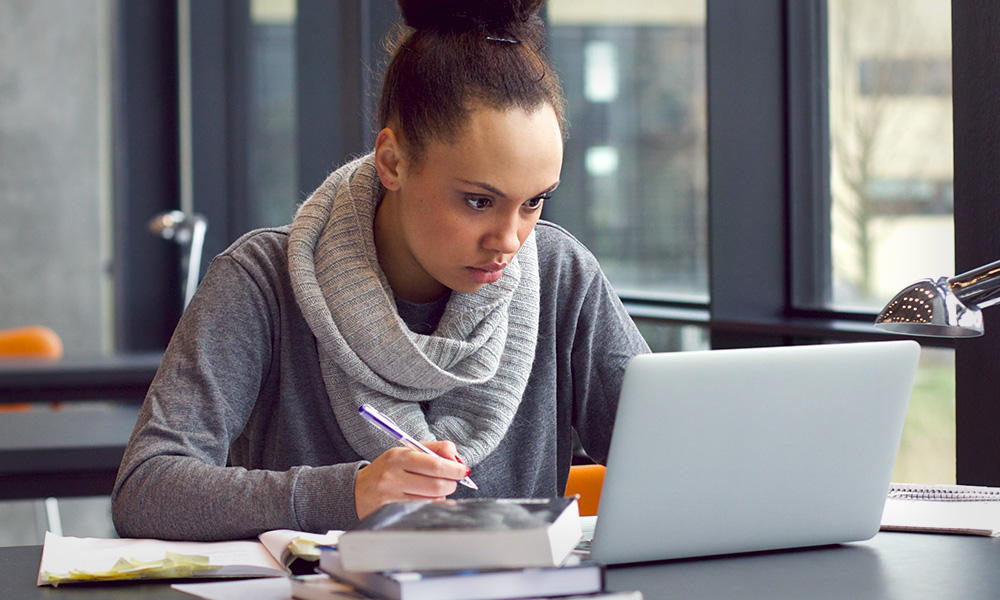 A woman sits at a desk, writing in a notebook with a pen while looking at an open laptop. The person is wearing a grey sweater and scarf. There are books and papers on the desk, and the background shows large windows with an outside view.