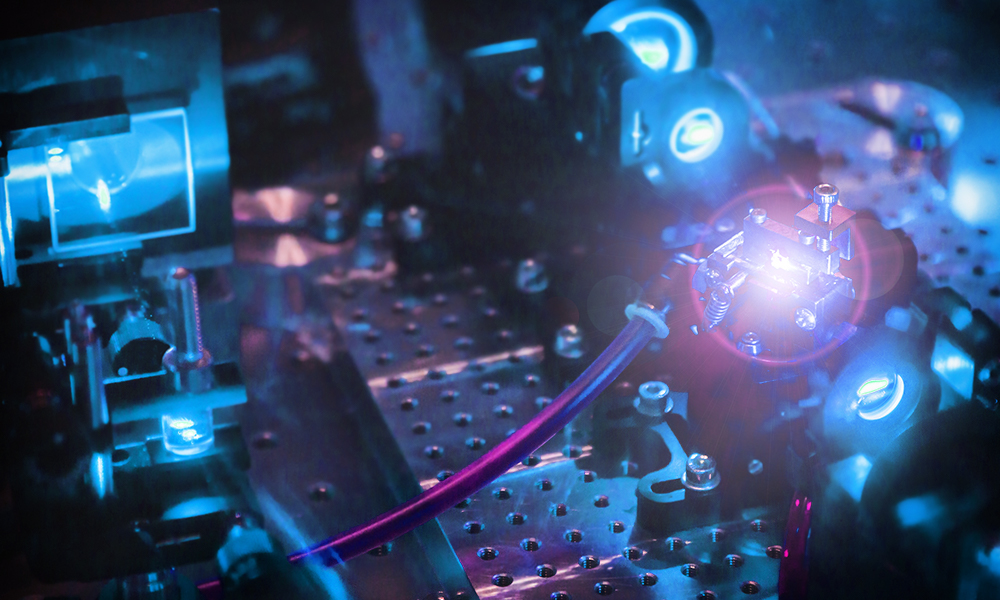 A close-up view of a high-tech laboratory setup involving various optical and electronic components, including several metallic parts, screws, and wires arranged on a perforated metal surface. Blue and purple lights illuminate the scene, and a prominent component in the center emits a bright blue light.