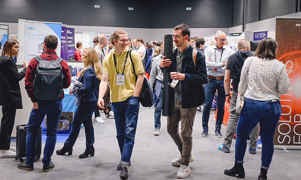 A busy exhibition hall with multiple people walking and conversing among various booths. Attendees are wearing conference badges and carrying bags or backpacks. Booths display banners with visible text such as “PIA Supercontinuum Fiber Lasers” and “SOLUTIONS” alongside colorful graphics. The setting appears professional, with bright lighting and a modern trade show environment.