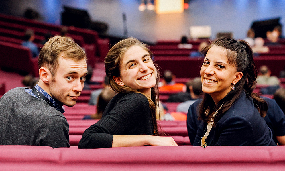 Three smiling attendees at SPIE Photonics Europe seated in a maroon auditorium, turned toward the camera during a conference or seminar, with other participants and a stage visible in the background.