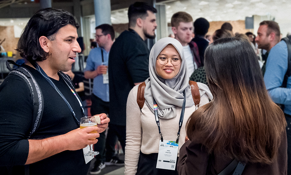 A group of conference attendees wearing name badges talk together in a bright networking area as they engage in conversation.