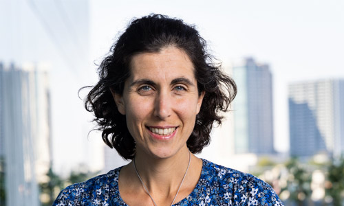Portrait of a person with short curly hair wearing a blue floral-patterned top and a necklace, standing outdoors with tall buildings and a clear sky in the background.