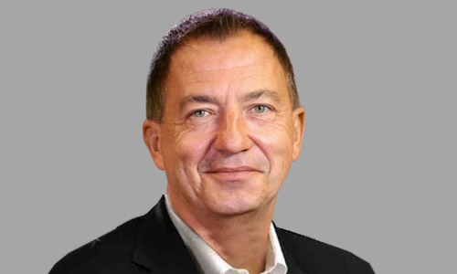 Head-and-shoulders portrait of a person in a dark blazer and white collared shirt on a neutral gray background.